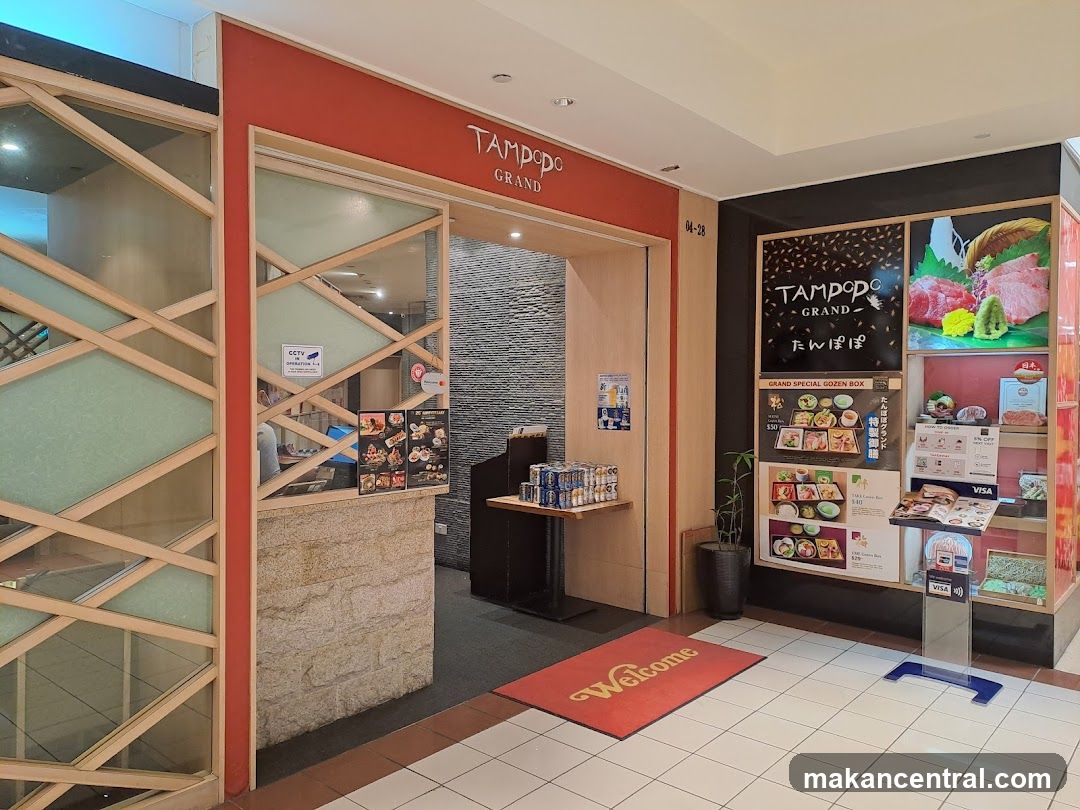 The exterior entrance of Tampopo Grand, a Japanese restaurant. The entrance features a red awning with the restaurant's name in white lettering. To the right of the entrance, there is a display board with images of food and prices. A red welcome mat is placed on the floor in front of the entrance.