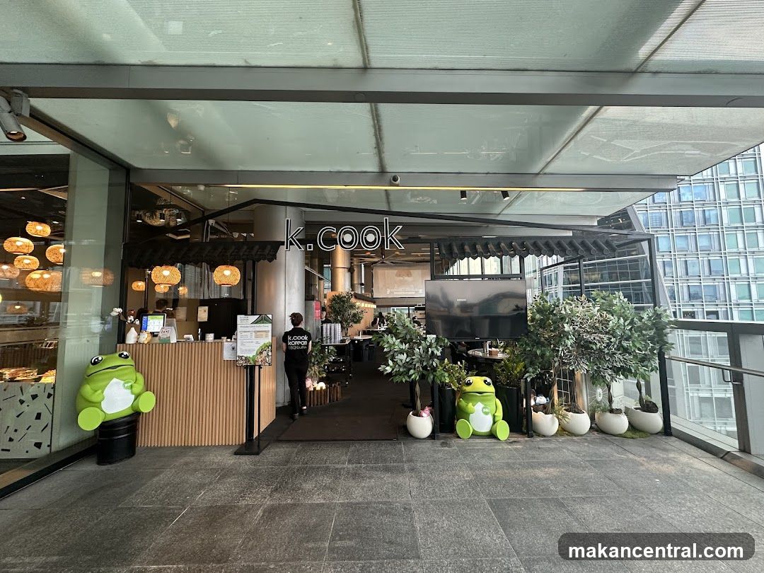 The exterior entrance of a Korean restaurant named K.cook. The restaurant has a modern facade with a prominent sign above the entrance. Inside, there are tables and chairs visible, along with decorative plants and lighting. Two large green frog statues are placed outside the entrance.