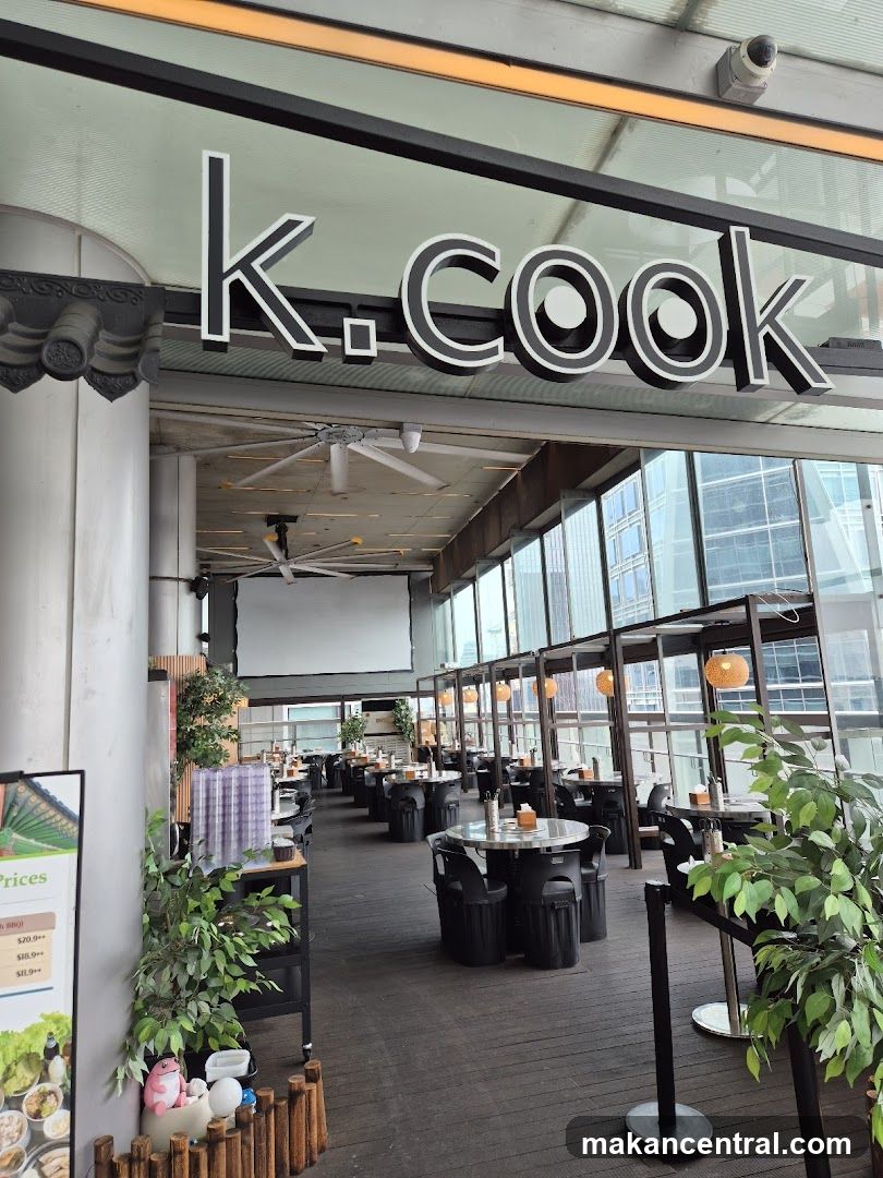The exterior of a restaurant with a large sign that reads "K. Cook". The restaurant has an outdoor seating area with many tables and chairs, and large windows overlooking a city skyline. There are plants and decorative elements around the seating area.