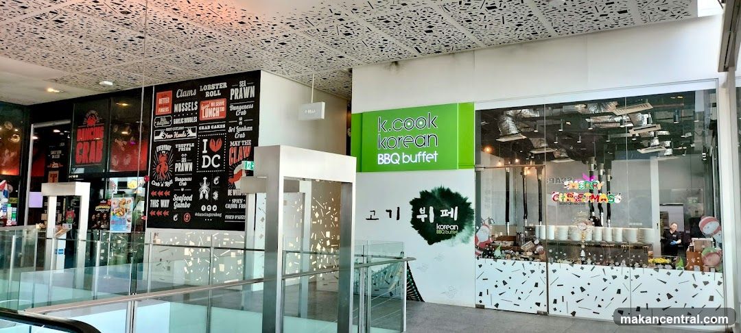 The exterior of a Korean BBQ buffet restaurant. A large green sign reads "k.cook korean BBQ buffet". Below this, a white wall features Korean text and a green graphic. The entrance is through glass doors, with a display of plates and Christmas decorations visible inside. To the left, a black wall with white text lists various seafood items and promotions for "Dancing Crab".