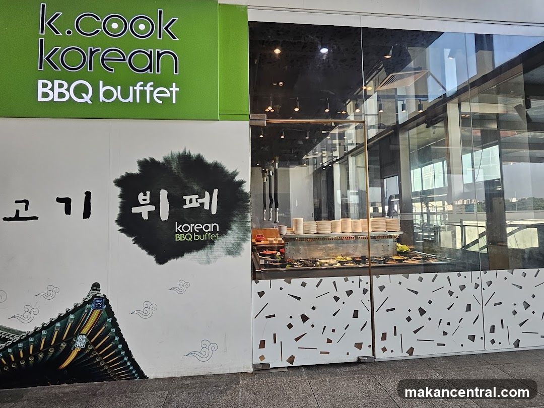 The exterior of a K.Cook Korean BBQ buffet restaurant. The signage above the entrance reads "k.cook korean BBQ buffet" in white lettering on a green background. To the left of the entrance, there is Korean text and a graphic of a traditional Korean roof. The entrance itself is a glass door with a glass facade, revealing a buffet setup inside with stacks of plates and various food dishes.