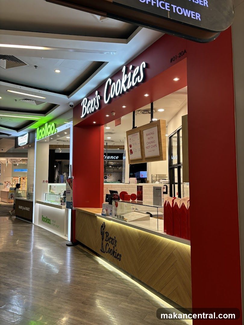 A brightly lit cookie shop with a red facade and a wooden counter. The shop's name, "Ben's Cookies," is displayed in large white lettering above the counter. There are two wooden signs hanging from the ceiling, one with text about baking and chunks, and another with a cookie graphic. The counter has a glass display case and various products.