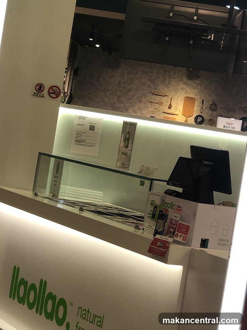 The image shows the counter area of a Llaollao store. The Llaollao logo is prominently displayed in green on the white counter. Behind the counter, there is a glass display case, a point-of-sale system with a monitor, and various promotional materials. A sign that says "BUY 10" is visible, along with a box featuring two cartoon ice cream cones and the text "natural frozen y". There are also signs indicating "No Pets Allowed" and "No Smoking". The overall impression is a clean and modern retail environment.