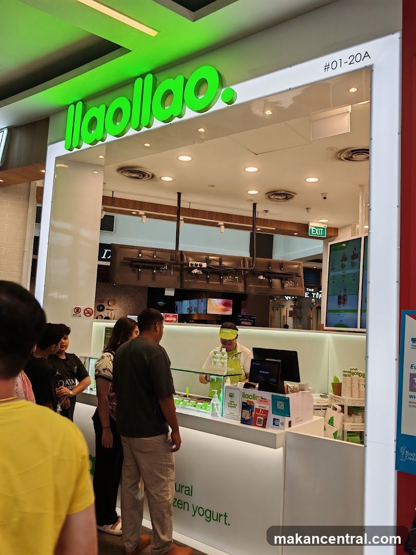 A brightly lit yogurt shop with the brand name "laollao." prominently displayed in green neon letters above the entrance. Customers are queuing to order from a staff member behind the counter. The counter displays various items and a screen. The shop's slogan "natural frozen yogurt." is visible on the lower part of the counter.