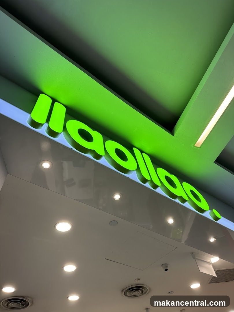 A bright green, illuminated sign with the text "llaollao." is mounted on a white facade. The sign is angled upwards, suggesting it is part of a storefront. The ceiling above is painted green and features recessed lighting and air vents.