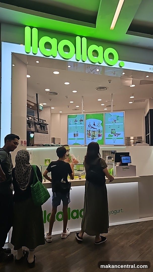 A group of people are standing in line at a llaollao frozen yogurt shop. The shop has a bright green sign with the name "llaollao." visible above the entrance. Inside, there are digital menus displaying various frozen yogurt options and toppings. A staff member is serving customers at the counter.