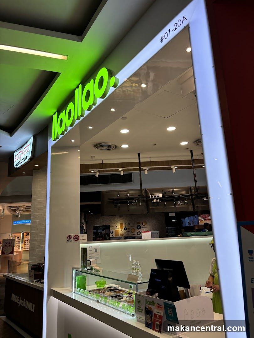 A brightly lit storefront for a frozen yogurt shop. The prominent green logo "llaollao" is visible above the entrance. The shop features a clean, modern design with white counters and illuminated panels. Inside, a display case shows various toppings for frozen yogurt, and a staff member in a green apron stands behind the counter.