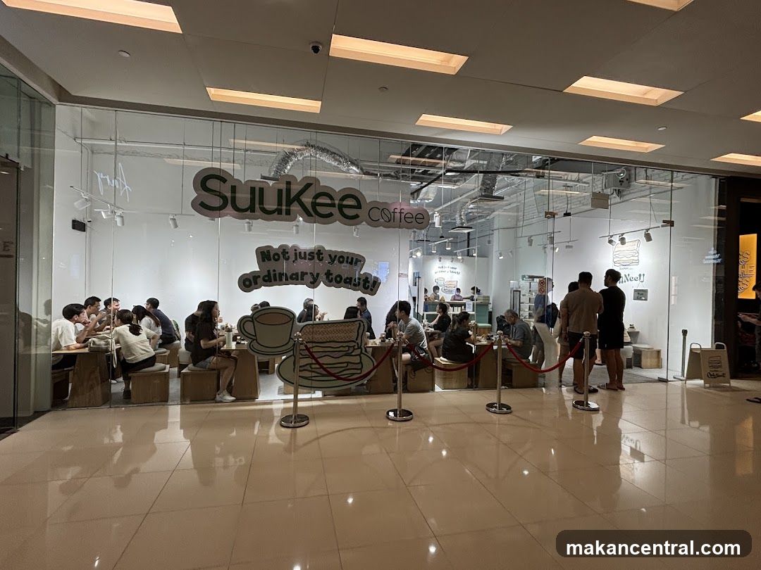A view of the interior of a cafe through its glass storefront. The cafe has a minimalist design with white walls and wooden furniture. Several patrons are seated at tables, enjoying their meals and drinks. The cafe's logo, "Suukee coffee," is prominently displayed on the glass, along with the slogan "Not just your ordinary toast!". A decorative display of a stack of toast and a cup of coffee is also visible.
