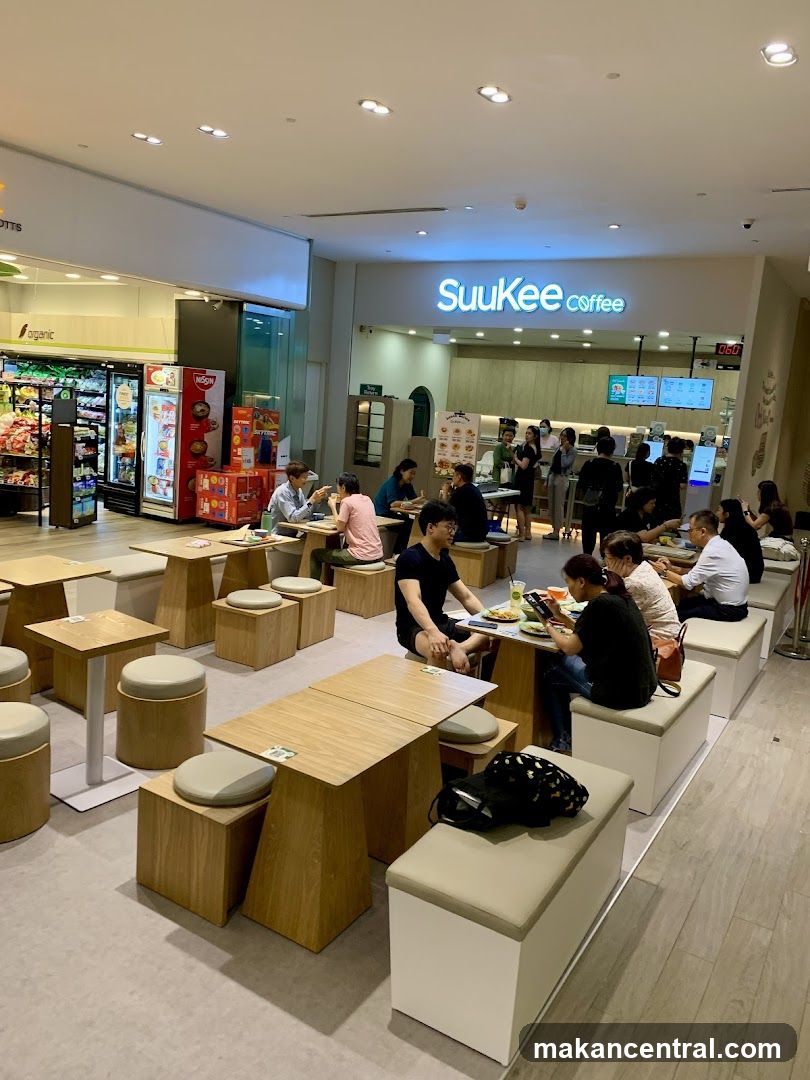The exterior of a coffee shop with a prominent "SuuKee coffee" sign. Inside, customers are seated at tables and benches, some eating and others looking at their phones. The shop appears to be located within a larger retail space, with a grocery store visible to the left.