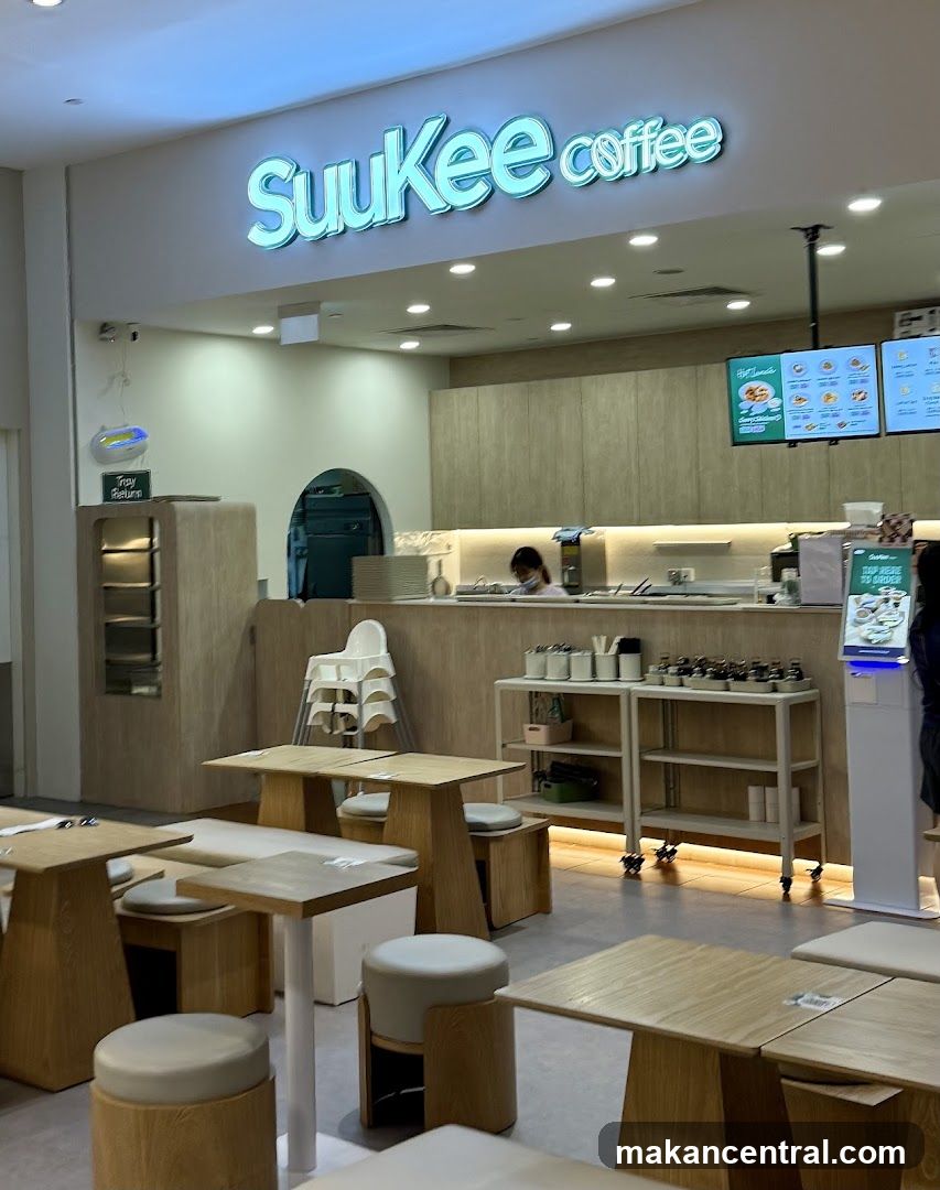 The exterior of a coffee shop with a prominent "SuuKee coffee" sign. The interior features light wood tables and seating, a counter with staff, and menu boards displayed on the wall.