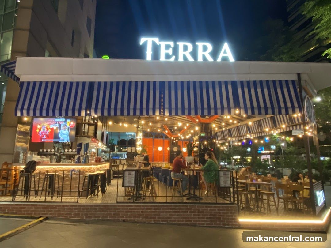 The exterior of a restaurant called Terra, with a blue and white striped awning and string lights. There is a bar area visible inside, with stools and a television screen. Several tables and chairs are set up outside, and people are dining. The restaurant has a modern, inviting ambiance.