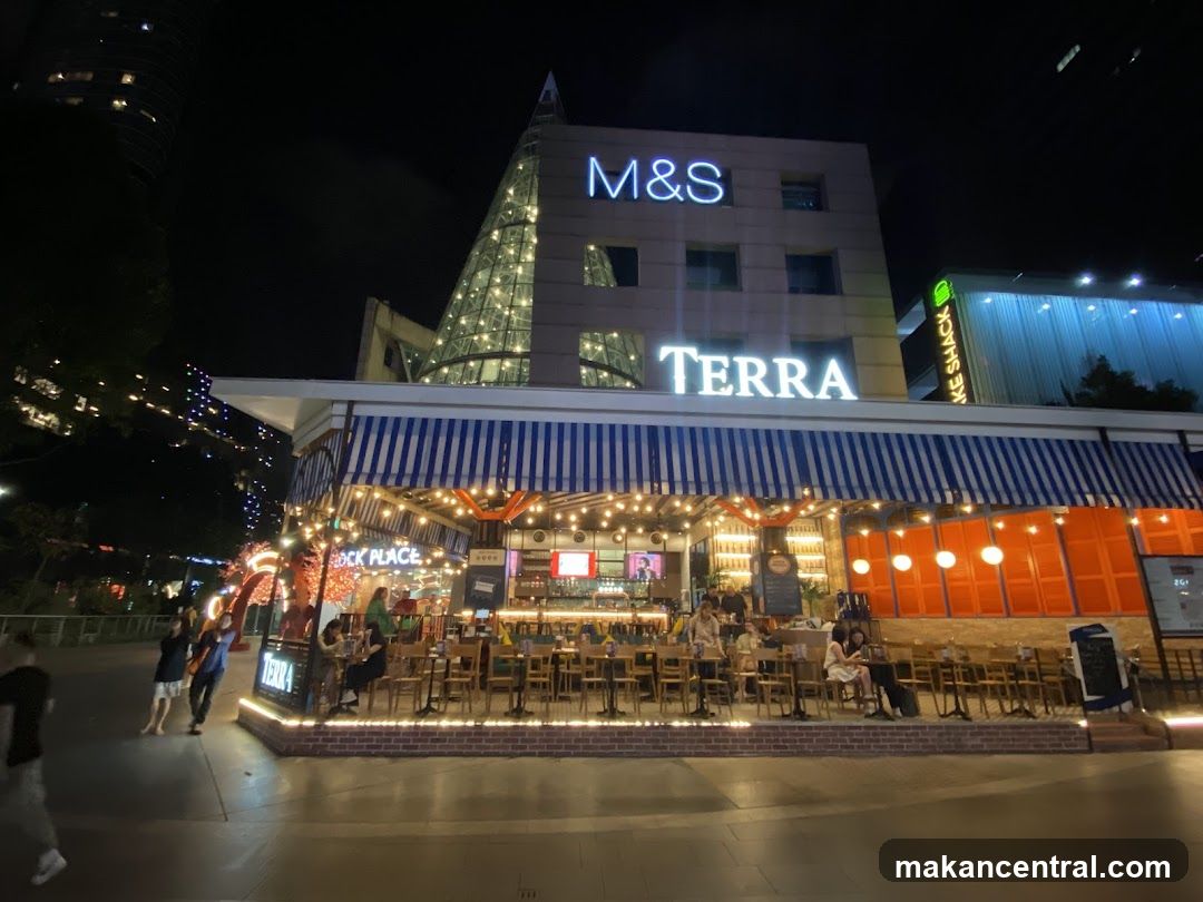 A night view of the Terra restaurant with outdoor seating. The restaurant has a blue and white striped awning and is lit with string lights. The M&S logo is visible on the building above.