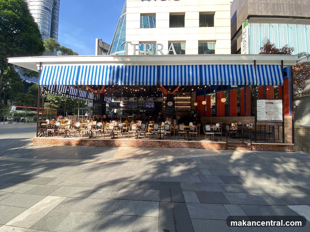 A street-level view of a restaurant with outdoor seating. The restaurant has a blue and white striped awning and tables and chairs set up on a brick patio. The building behind the awning has the word "TERRA" visible in large letters.