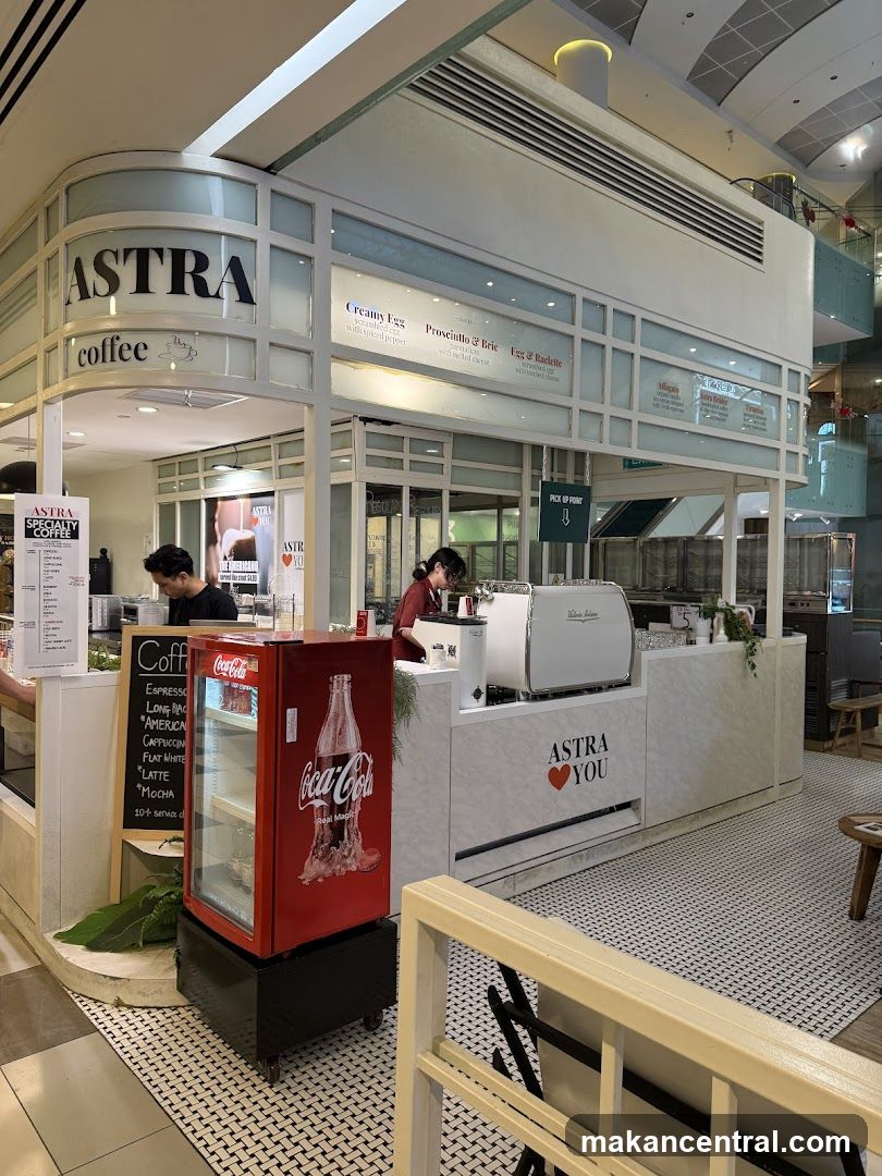 The exterior of a modern coffee shop with the name ASTRA prominently displayed. The shop features a clean white design with glass panels. A red Coca-Cola refrigerator is visible to the left, and a coffee machine and counter are to the right. A menu board with coffee and food items is also visible.