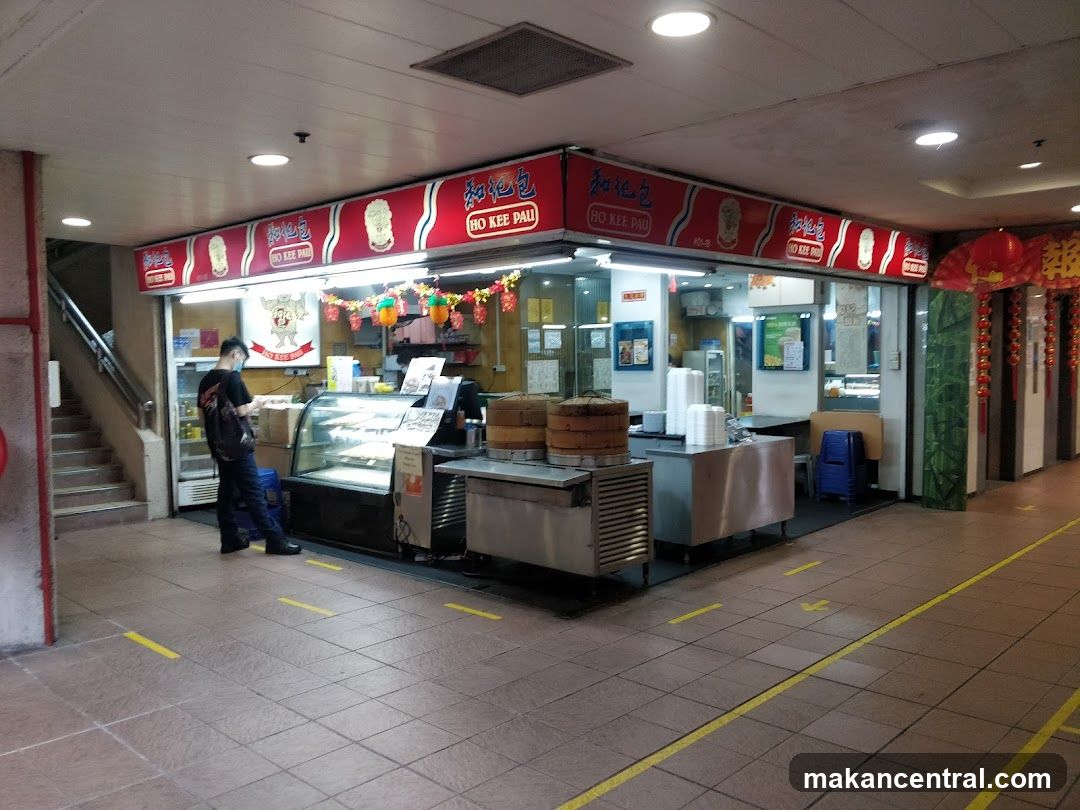 A view of the exterior of a food stall named Ho Kee Pau. The stall has a red awning with the name in Chinese characters and English. There is a display case with food items and steaming baskets. A person is standing in front of the stall.