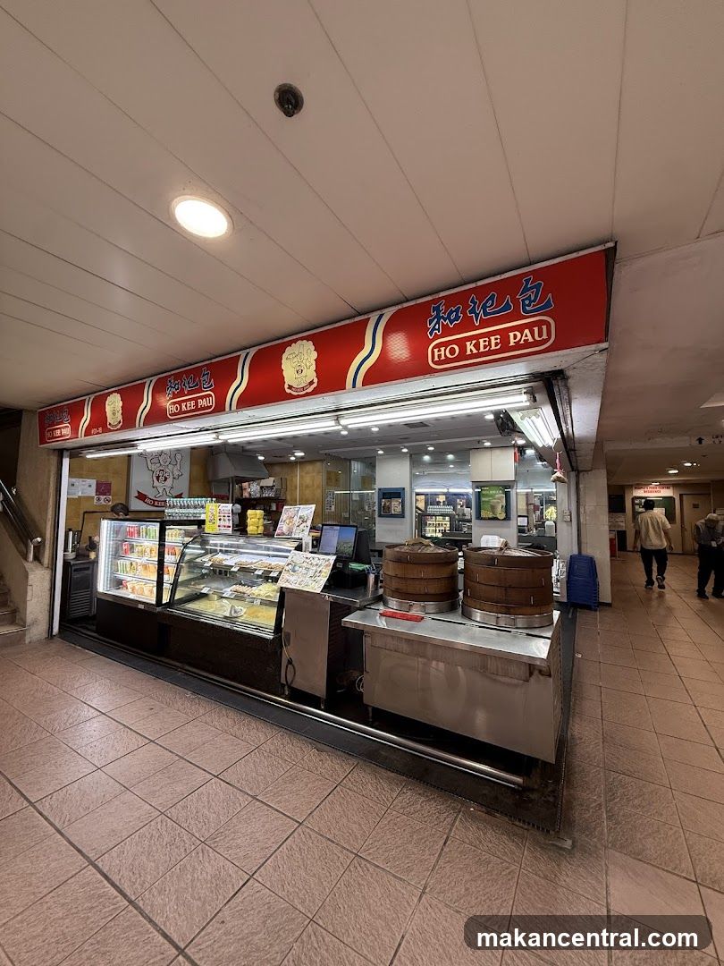 A view of the exterior of a food stall with a red awning displaying the name "HO KEE PAU" in English and Chinese characters. Below the awning, a display case shows various food items, and two large bamboo steamers are visible. The stall is situated in a tiled walkway with other shops and people in the background.