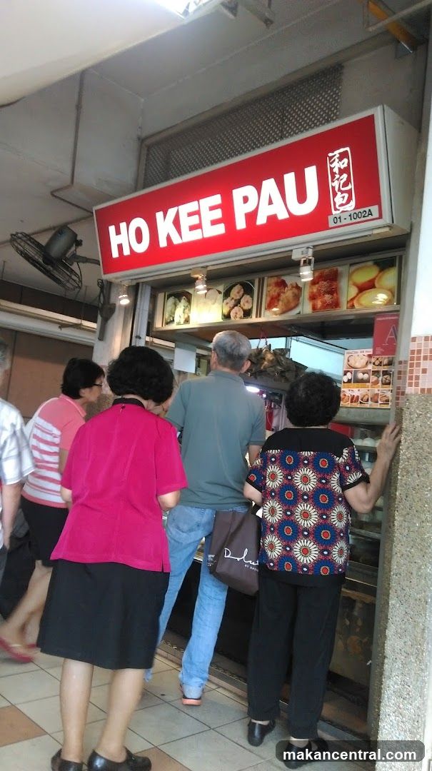 A bright red sign with white lettering that reads "HO KEE PAU" is prominently displayed above a food stall. Below the sign, a display case shows various food items, and a menu board with pictures of food is visible. Several people are standing in front of the stall, appearing to be customers.