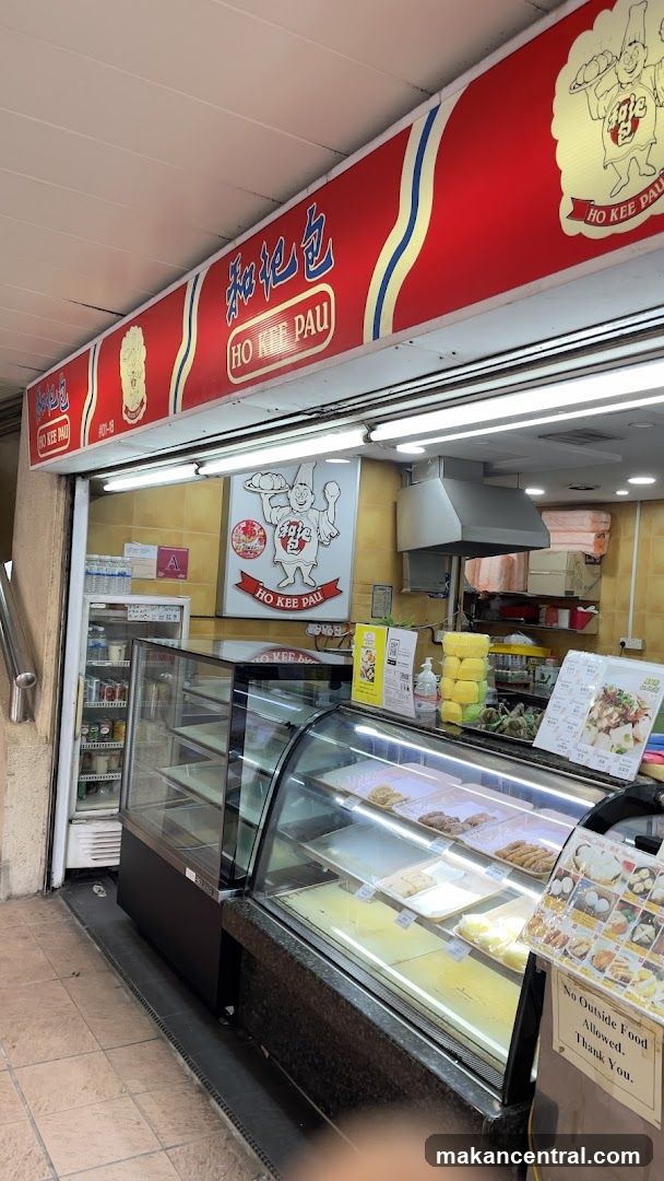 The exterior of a food establishment with a prominent red sign displaying "HO KEE PAU" in both Chinese characters and English. Below the sign, a display case showcases various food items. A refrigerator is visible to the left, stocked with drinks. A sign on the display case reads "No Outside Food Allowed. Thank You."