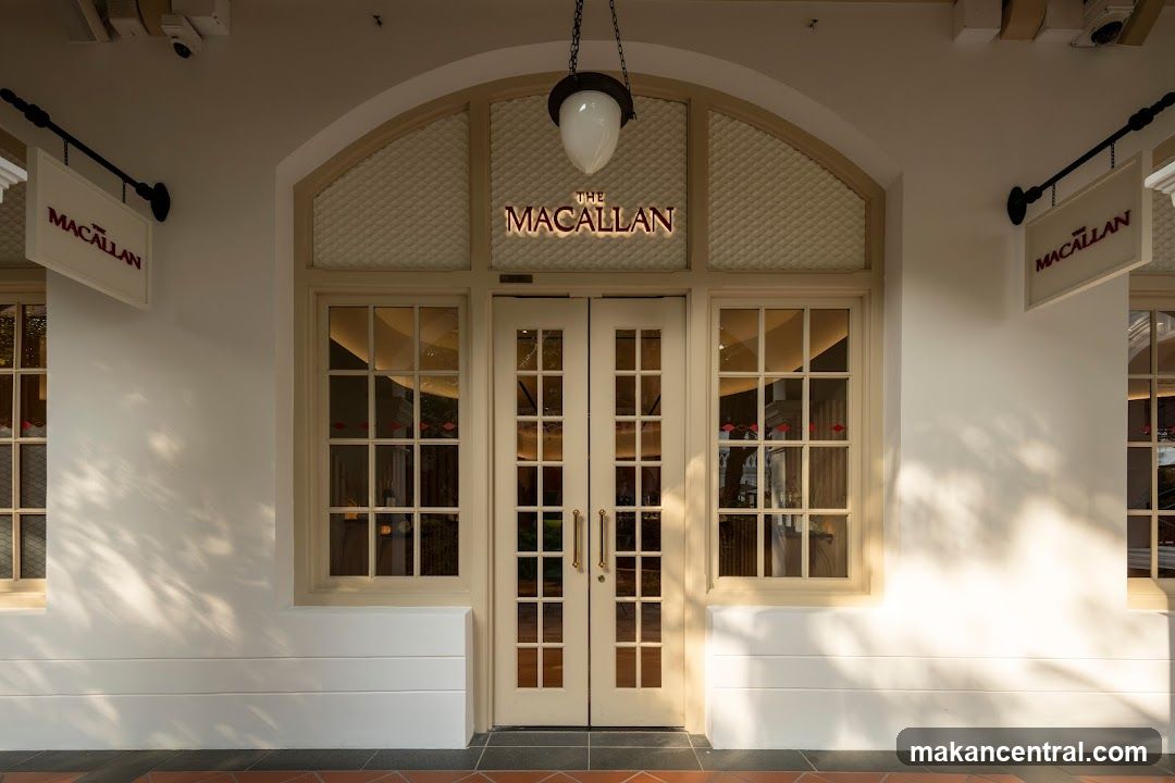 A well-lit storefront with double doors and large windows. Two signs with the name "THE MACALLAN" are visible, one above the doors and another hanging to the left. The building is painted white with cream-colored trim around the windows and doors.