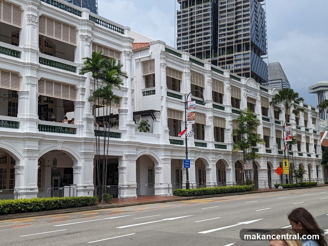 A white colonial-style building with multiple stories and arched entrances. The word "RAFFLES" is visible in large letters above the second story. Palm trees are planted in front of the building. Modern skyscrapers are visible in the background.