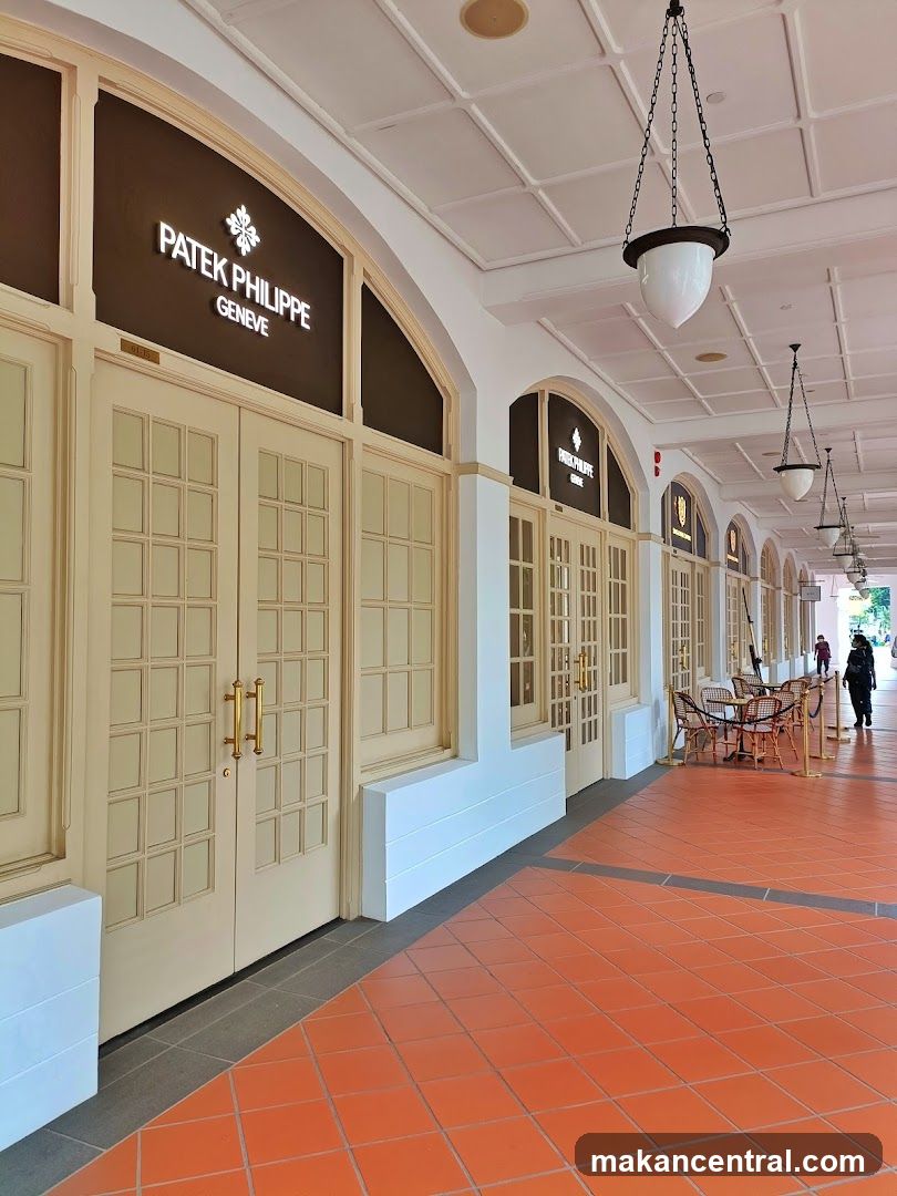 A row of arched doorways with cream-colored double doors and gold handles. Above each doorway, a dark brown panel displays the Patek Philippe Geneve logo and name in white lettering. The storefront is part of a covered walkway with a white tiled ceiling and hanging pendant lights. The walkway has an orange tiled floor. In the distance, tables and chairs are set up outside one of the establishments, and a few people are walking along the path.