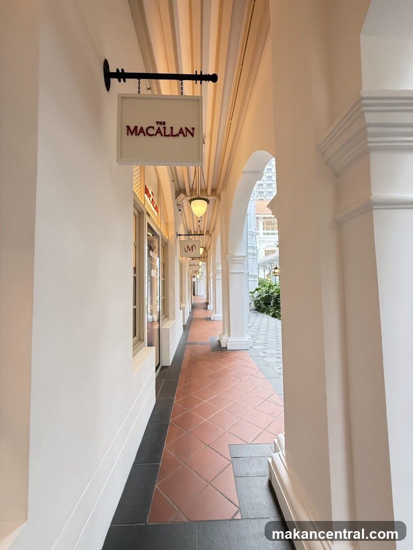 A view down a covered walkway with shops on the left. A sign for "THE MACALLAN" hangs prominently on the left wall. Further down the walkway, other signs and shop windows are visible. The walkway has tiled flooring and arched openings on the right leading to an outdoor area.