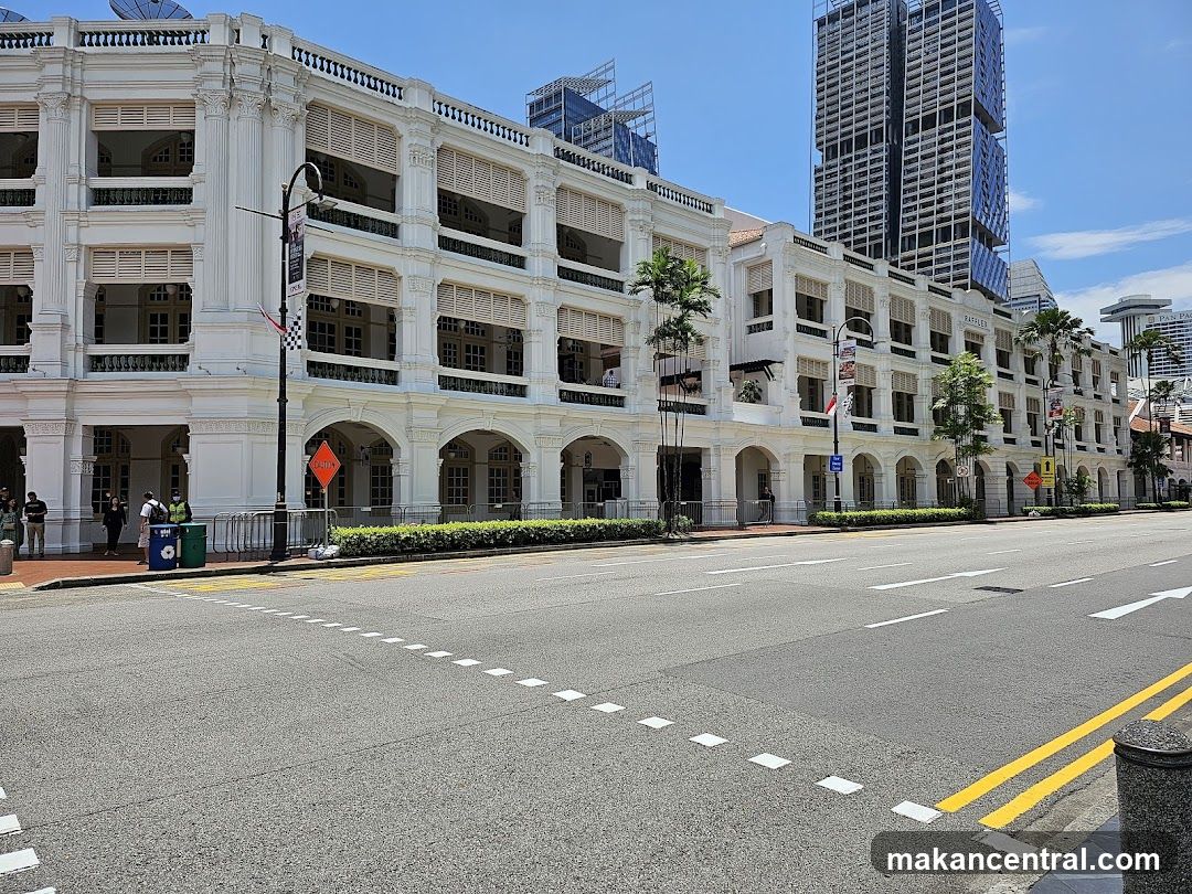 A wide shot of the historic Raffles Hotel building on a sunny day. The white, multi-story colonial-style building features arched walkways on the ground floor and shuttered windows on the upper levels. Palm trees line the sidewalk in front of the hotel. Modern skyscrapers are visible in the background. Several people are standing on the sidewalk near the entrance.