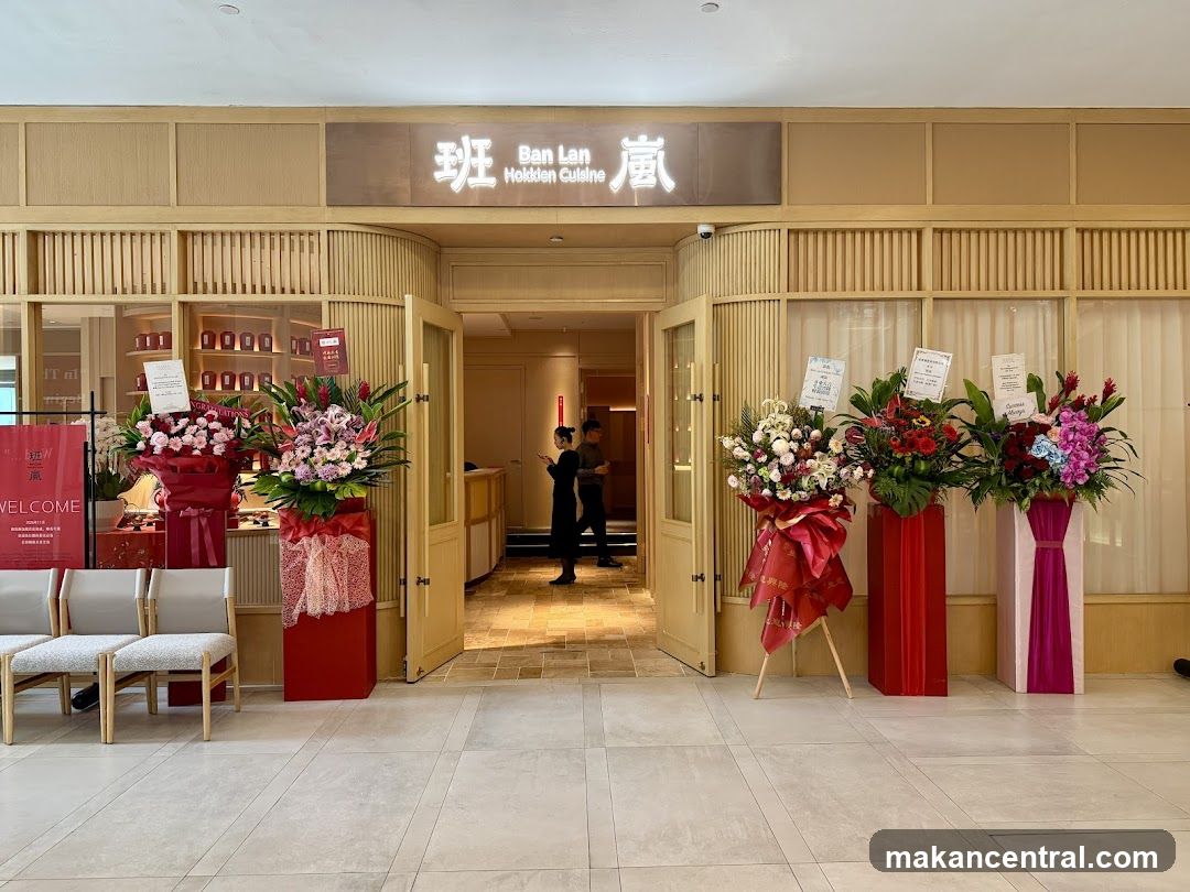 The image shows the front entrance of a restaurant with wooden paneling and a sign displaying '班 Ban Lan 廬' above the doorway. The entrance is flanked by red floral arrangements on stands, suggesting an opening or celebration. Inside, two people are visible near the reception area. The floor is tiled, and there are chairs to the left.