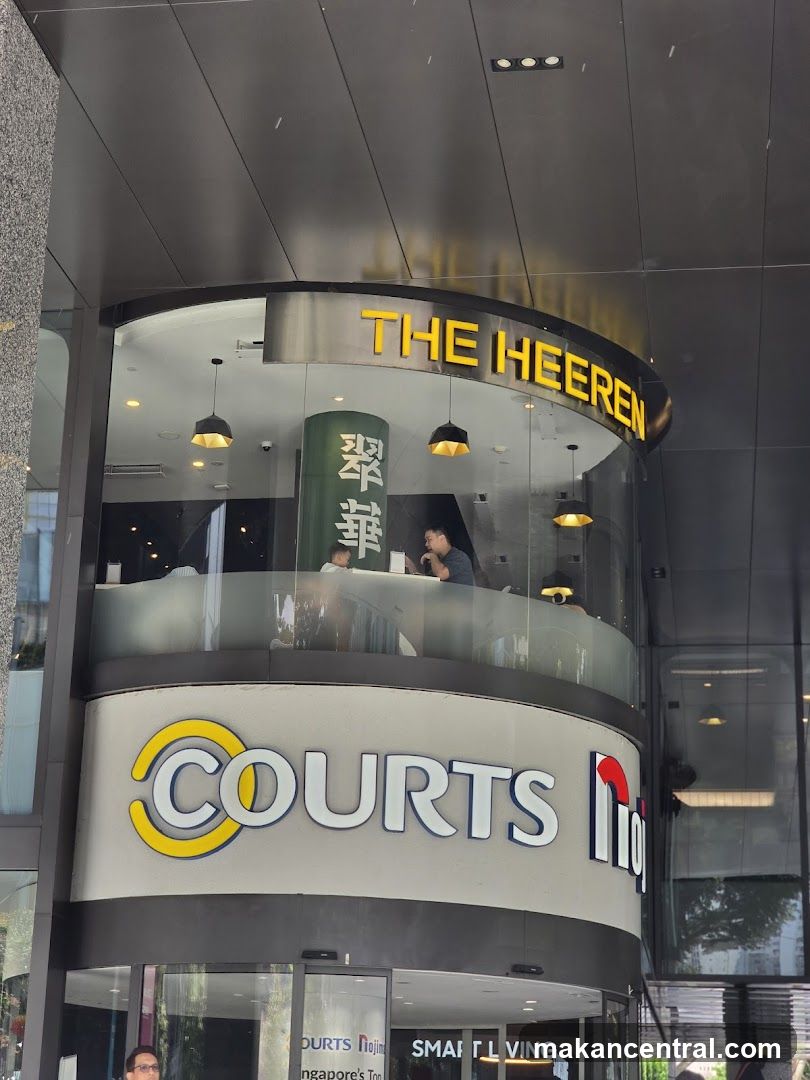 Exterior view of a modern curved glass facade with 'THE HEEREN' in yellow lettering above the second level and 'COURTS' logo below it. Visible through the glass are interior lighting fixtures and patrons seated inside.
