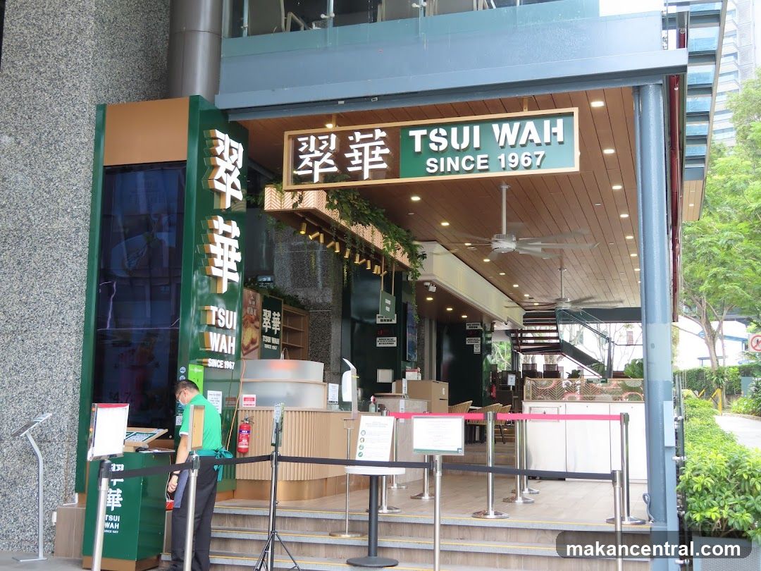 Exterior view of TSUI WAH restaurant entrance with green signage displaying "翠華" and "SINCE 1967" above the doorway. A staff member in a green shirt is at the counter area. The interior features wooden ceilings, hanging plants, and red stanchion barriers leading to the dining area.
