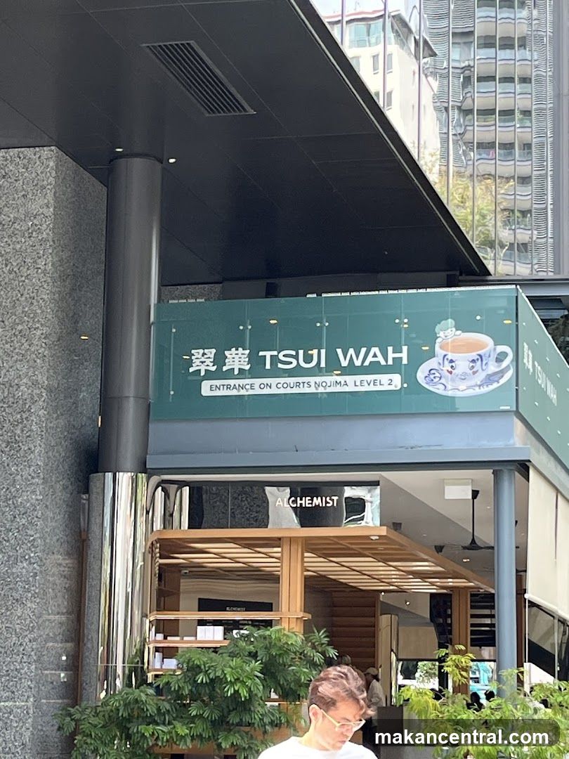 Exterior storefront with green sign displaying "翠華 TSUI WAH" and "ENTRANCE ON COURTS NQJIMA LEVEL 2". Below is an open wooden-framed entrance with a visible interior featuring the word "ALCHEMIST" on a black panel. Green plants are placed at the base of the entrance. A person wearing glasses walks past in the foreground.