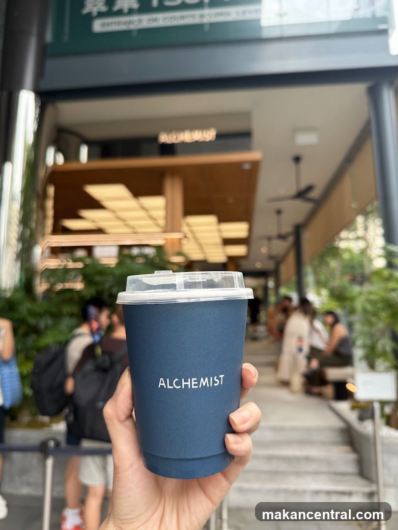 A hand holds a blue Alchemist branded cup in front of the storefront entrance. The shop's name is visible on a sign above the doorway, with modern architectural elements including wooden beams and recessed lighting inside. People are seen sitting outside and walking by, suggesting an active outdoor seating area.