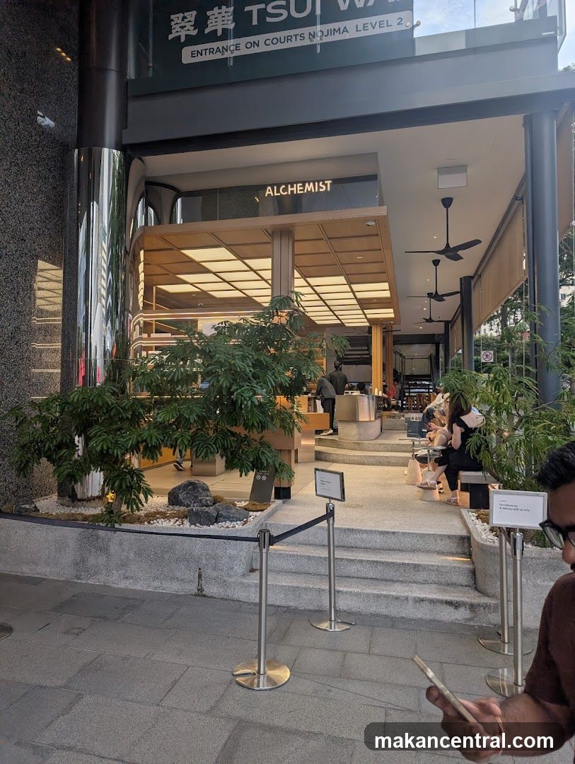Exterior view of a modern building entrance with glass facade and wooden accents. Signage above reads '翠華 TSU' and 'ENTRANCE ON COURTS NOJIMA LEVEL 2'. Inside, the word 'ALCHEMYST' is illuminated on a sign above the main counter area. A small landscaped garden with rocks and plants sits at the base of the steps leading into the shop. Several people are seated inside or walking through the space. The interior features warm lighting, ceiling fans, and open-plan design.