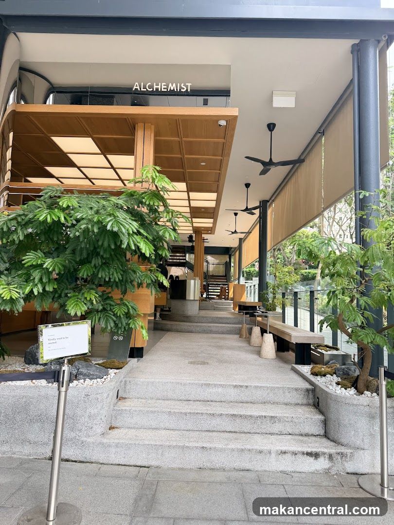 Exterior view of a modern building with wooden overhang and signage reading 'ALCHEMIST' above the entrance. Steps lead up to a covered walkway with benches, plants in stone planters on either side, and ceiling fans overhead. The facade features glass railings and natural lighting from skylights.