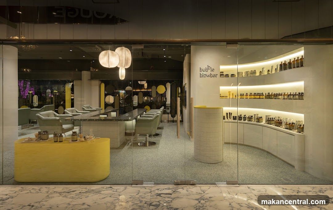 Glass storefront with modern design, featuring a curved white wall displaying shelves of products under LED lighting. The shop name 'bubble blowbar' is printed on the wall in black lowercase letters. A bright yellow cylindrical counter sits near the entrance, and inside, salon chairs and a central reception desk are visible. Above, spherical pendant lights hang from the ceiling. The floor outside is marble, and the interior has a speckled gray carpet.