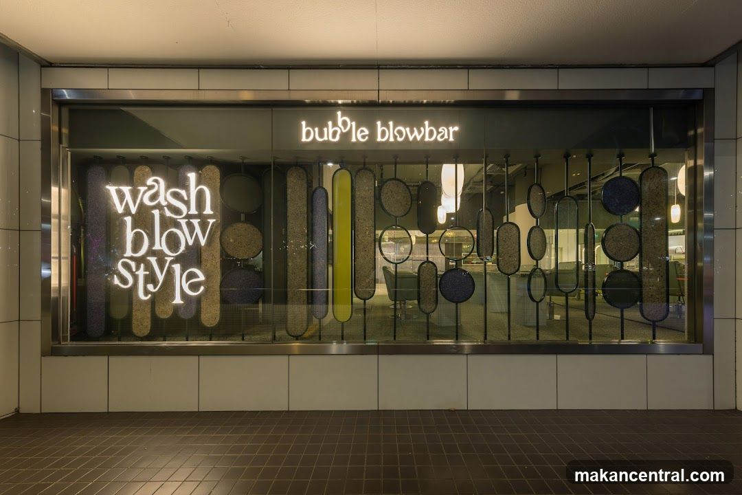 Storefront with illuminated sign reading 'bubble blowbar' above a large glass window displaying the phrase 'wash blow style' in glowing letters. The window features decorative vertical panels of varying shapes and textures, including circles and ovals, some with glittery or textured surfaces. Inside, salon chairs and lighting fixtures are partially visible through the glass. The exterior has a tiled floor and metallic frame around the window.