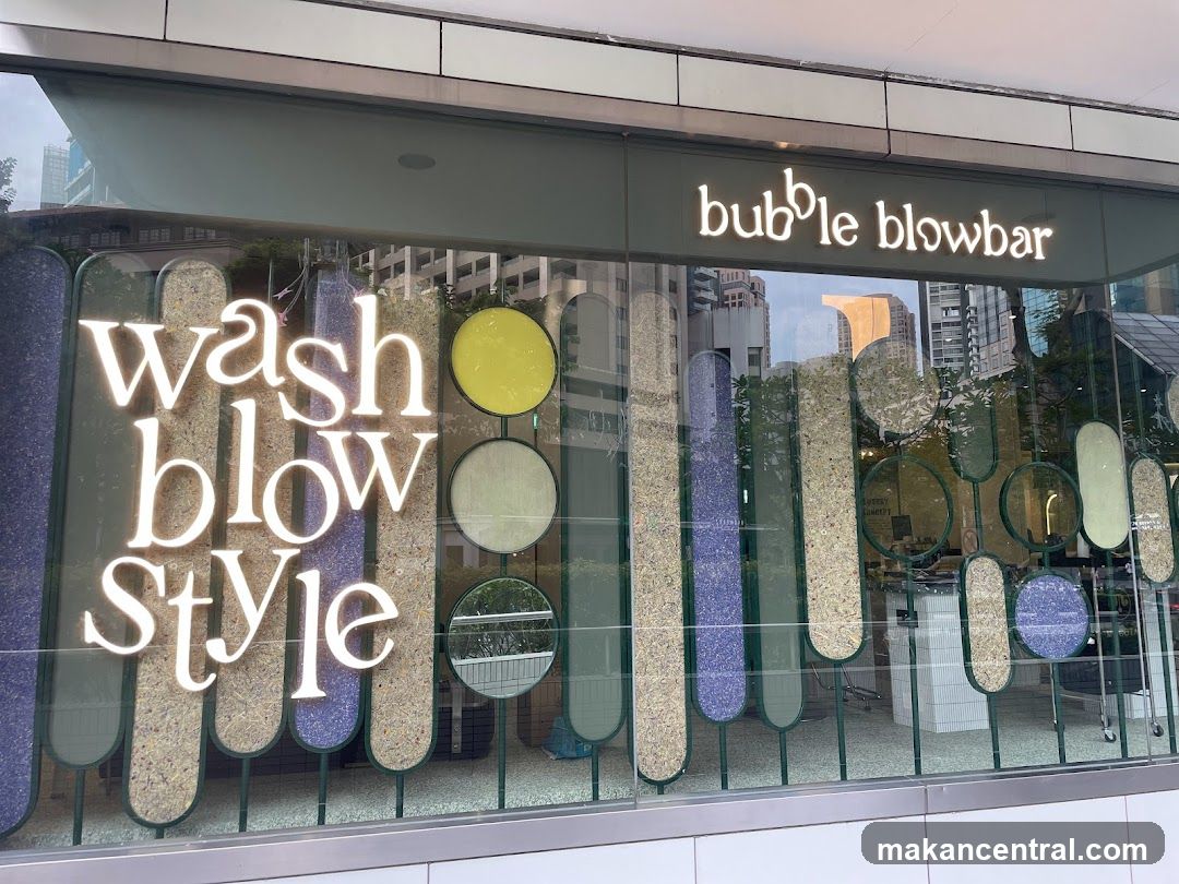 Modern glass facade with illuminated signage reading 'bubble blowbar' above the window and 'wash blow Style' in large white letters on the left side of the display. The window features decorative vertical panels with glittery textures in purple, beige, and blue tones, interspersed with circular cutouts in yellow, green, and blue. Reflections of surrounding buildings and trees are visible in the glass. No menu or printed materials are present.