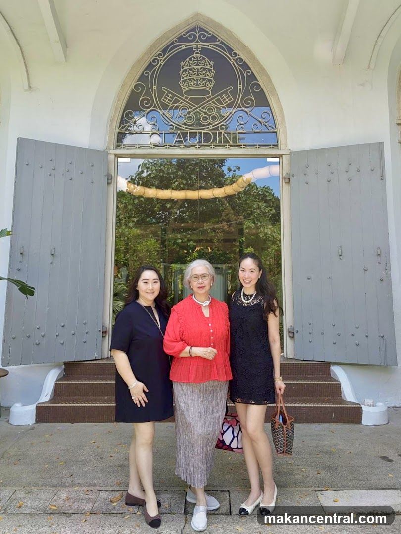Three women posing in front of a building entrance with arched glass door featuring the name 'CLAUDINE' and an ornate metal design above it. The doors are open, revealing greenery inside. The facade is white with grey shutters.