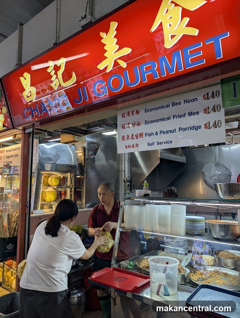 Red signboard with yellow and blue lettering displaying '昌記美食' and 'CHANG JI GOURMET'. Visible interior of food stall with stainless steel counter, cooking area, and staff interacting. Self-service setup indicated by signage.