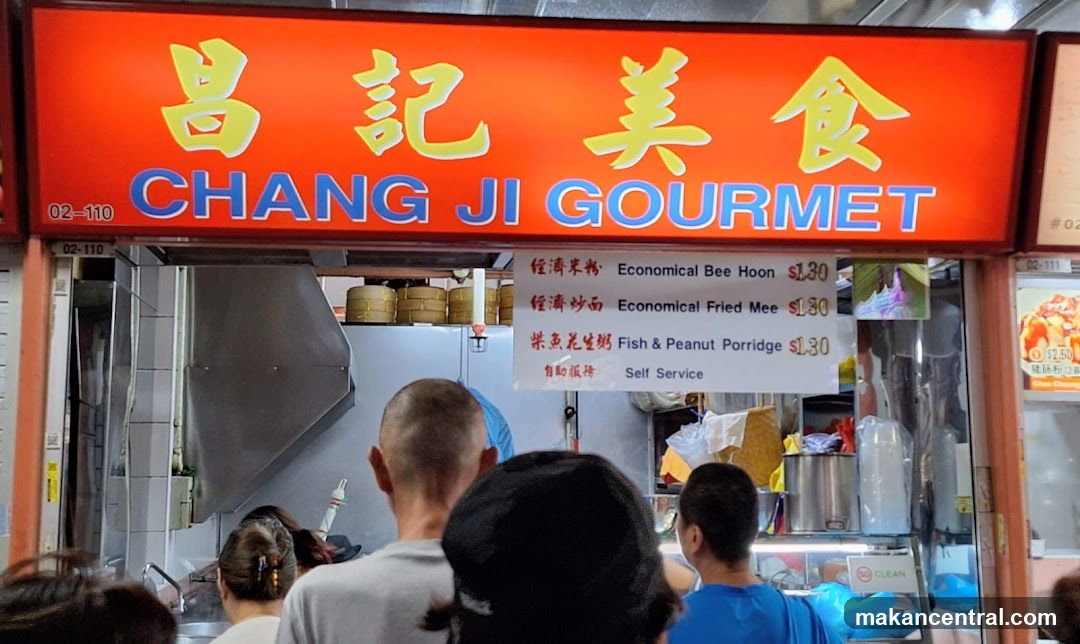 Exterior storefront with bright red sign featuring yellow Chinese characters '昌記美食' and blue English text 'CHANG JI GOURMET'. Below the main sign, a white menu board lists three items: 'Economical Bee Hoon', 'Economical Fried Mee', and 'Fish & Peanut Porridge', each priced at $1.30. The shop appears busy with customers visible from behind, facing the counter. A stainless steel vent hood is visible inside the kitchen area.