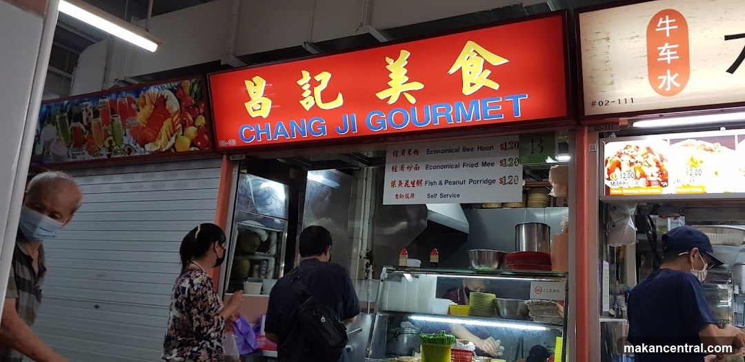Red signboard with yellow Chinese characters '昌記美食' and blue English text 'CHANG JI GOURMET'. Shopfront shows interior counter, staff preparing food, customers wearing masks. Adjacent stall has a vertical red sign with white Chinese characters '牛车水'.