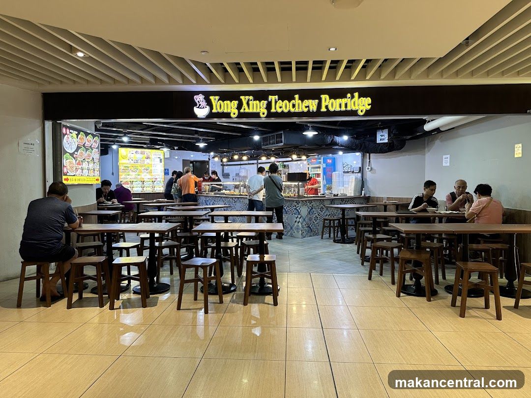 A wide shot of the Yong King Teochew Porridge shop front. The shop has a large yellow sign with the name of the shop in black lettering. Inside the shop, there are several tables and chairs set up for customers. Some customers are seated at the tables, eating. The counter area is visible, with food displayed. There are also menu boards visible.