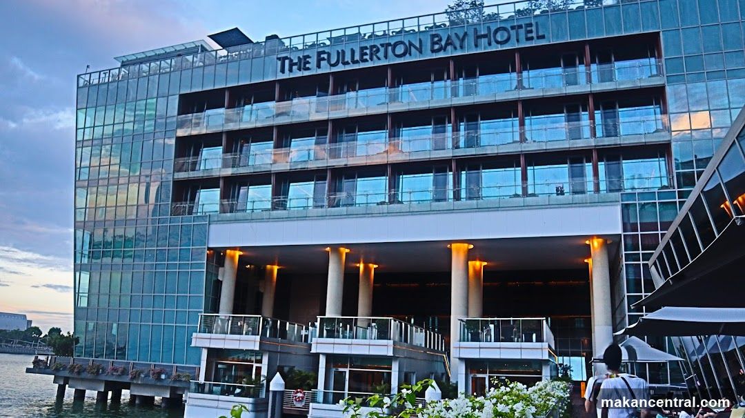 A modern glass-fronted hotel building with balconies and a rooftop sign that reads "THE FULLERTON BAY HOTEL". The hotel is situated on the waterfront, with reflections of the sky and surrounding buildings visible on its facade. The entrance area features illuminated columns and outdoor seating areas with umbrellas.