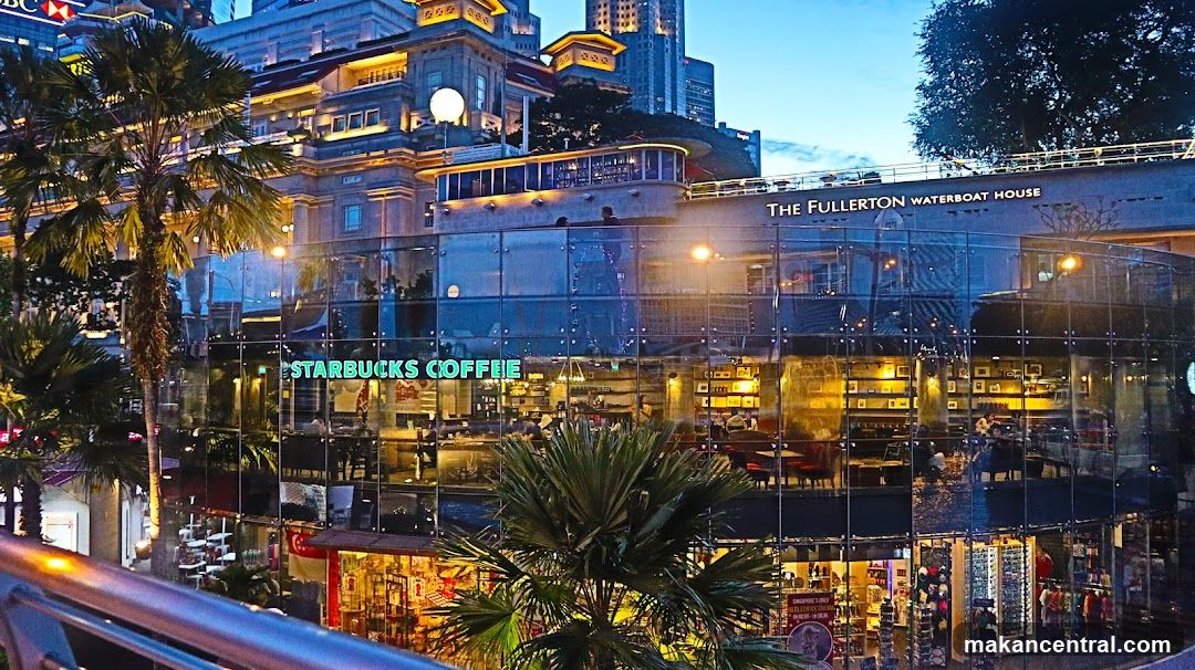 A modern glass-fronted Starbucks Coffee shop is visible on the ground floor of The Fullerton Waterboat House building. The Starbucks logo is prominently displayed on the glass facade. Inside, the warm lighting illuminates the seating area and shelves stocked with merchandise. The upper floors of the historic Fullerton Waterboat House building are visible above, with architectural details and lights. Palm trees frame the scene, and the sky is a deep twilight blue.