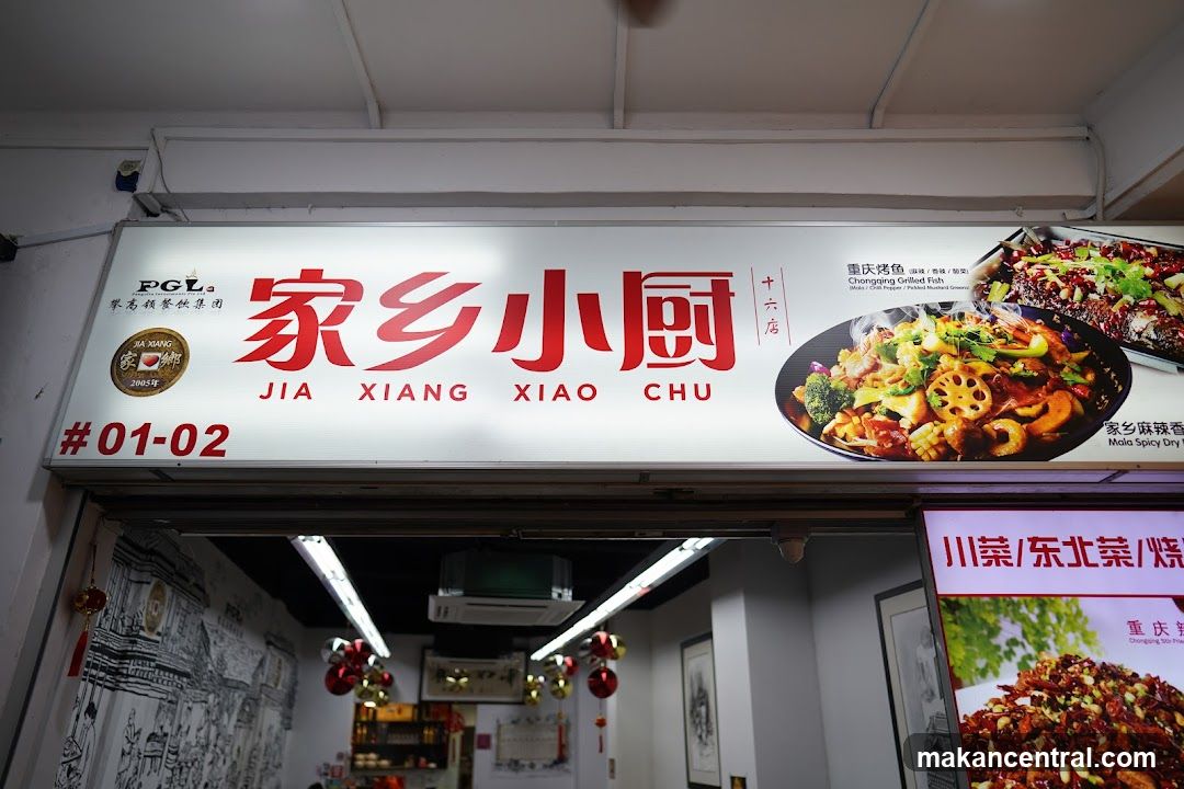 A brightly lit sign above a storefront displays the name "JIA XIANG XIAO CHU" in both Chinese and English. The sign also features images of two dishes: Chongqing Grilled Fish and Mala Spicy Dry. Below the main sign, a smaller illuminated sign shows images of food and text in Chinese, including "Chuan Cai / Dong Bei Cai / Shao Kao". The storefront entrance is visible below the signs, with a mural on the interior wall.