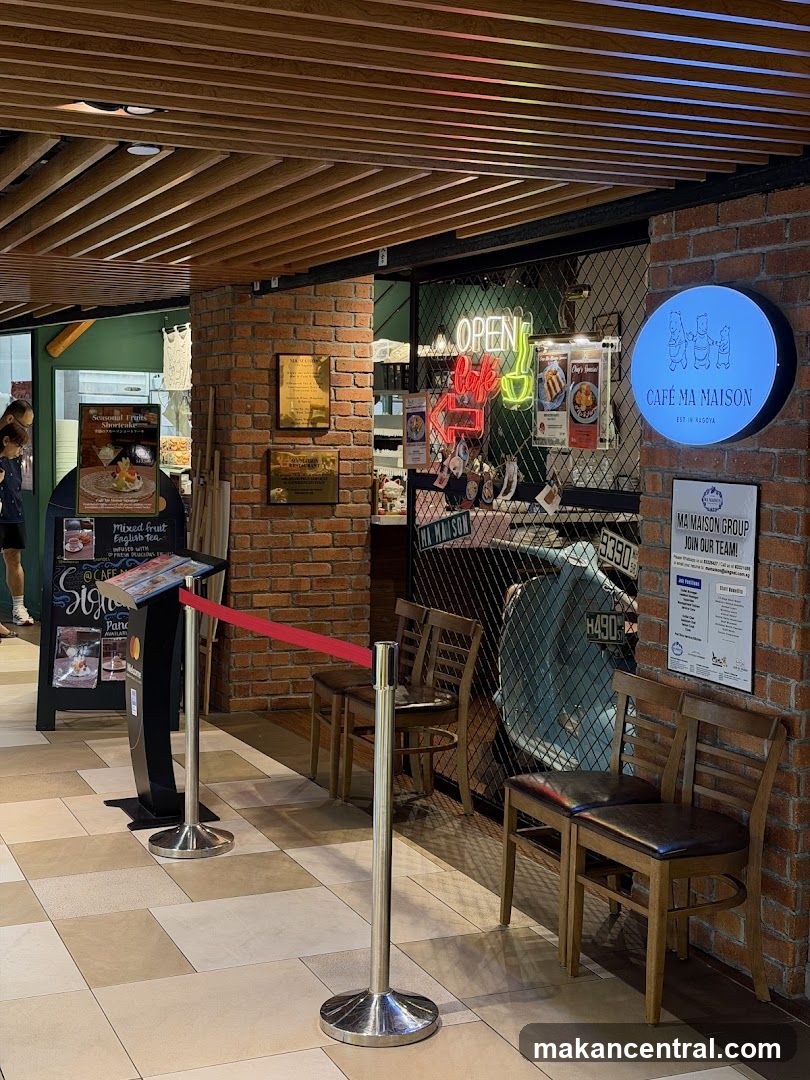 The exterior of a cafe with a brick facade. A circular blue sign with the cafe's name and logo is visible. Neon signs inside read "OPEN" and "cafe". A menu board is displayed near the entrance, and chairs are set up for waiting customers.