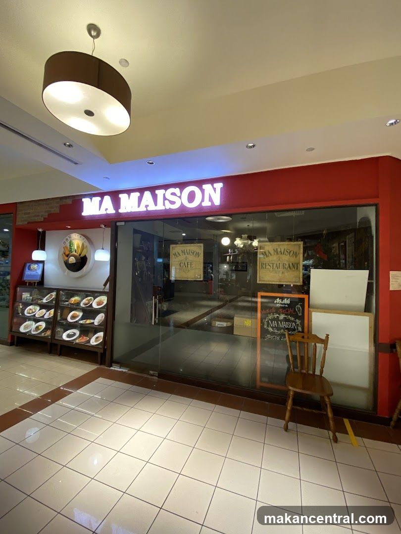 The exterior of a restaurant called Ma Maison. The restaurant has a large glass storefront with a prominent illuminated sign above that reads "MA MAISON". Inside, there are displays of food and menus. A wooden chair is placed outside the entrance.