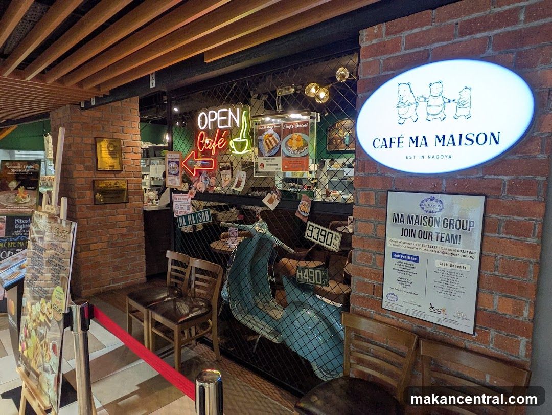 The exterior of a cafe with a prominent oval sign displaying "CAFÉ MA MAISON EST IN NAGOYA" and three cartoon bears. A "JOIN OUR TEAM!" poster is also visible on the brick wall. Inside, a neon "OPEN Café" sign and various food images are displayed.