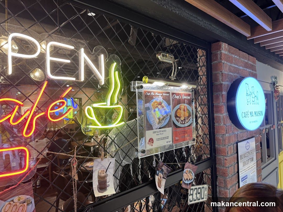 A neon sign that says "OPEN" and "café" is visible on the glass storefront of a cafe. A circular sign with the cafe's logo and name "CAFÉ MA MAISON" is mounted on a brick wall. Two menu boards are displayed behind a metal grate.