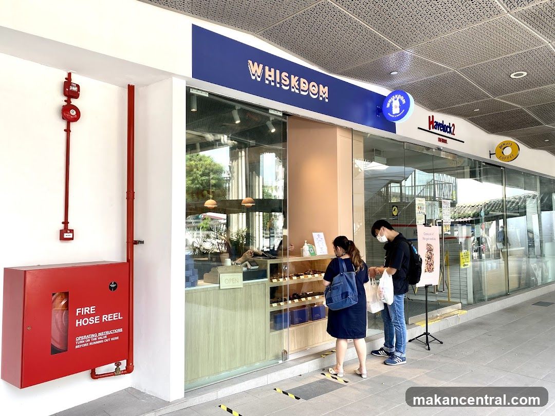 The exterior of a bakery called Whiskdom, with a large blue sign above the entrance. Two customers are standing at the counter, looking at the pastries. A fire hose reel is mounted on the wall to the left of the entrance.